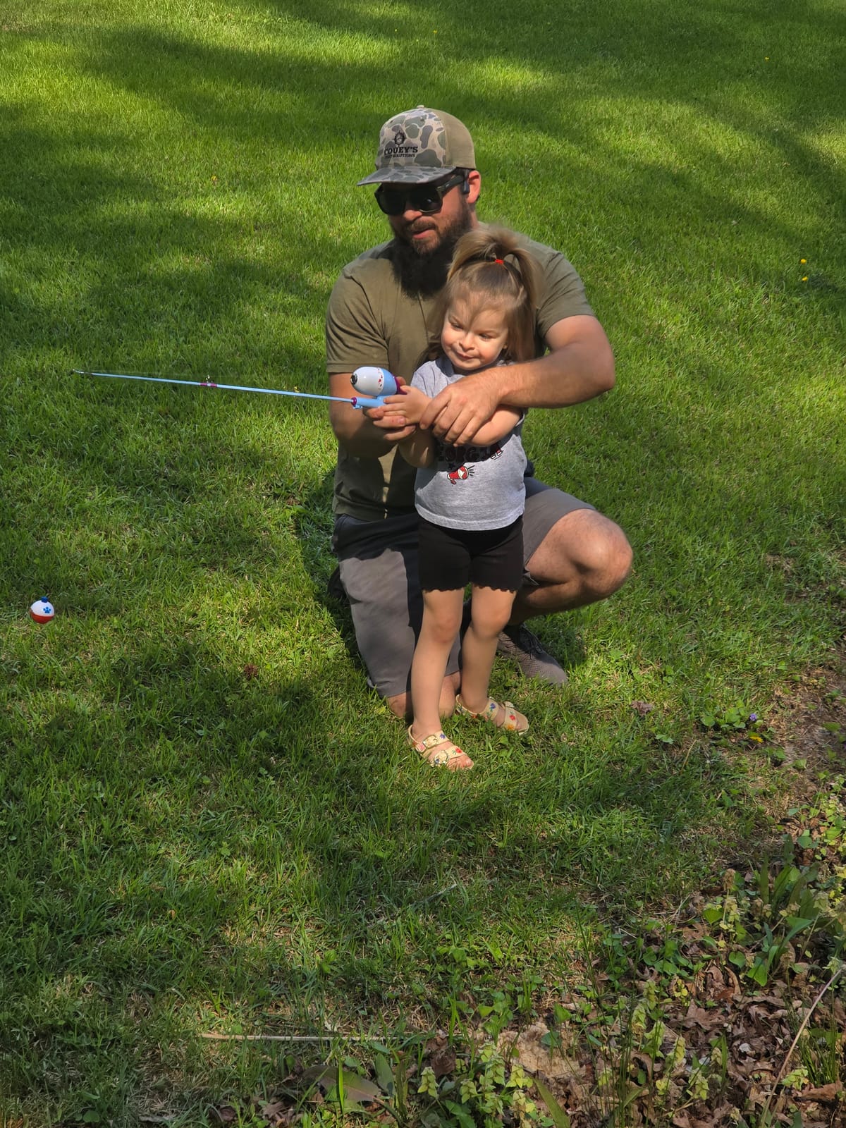 Brandon Turley crouching in the grass, helping a little girl hold a fishing rod